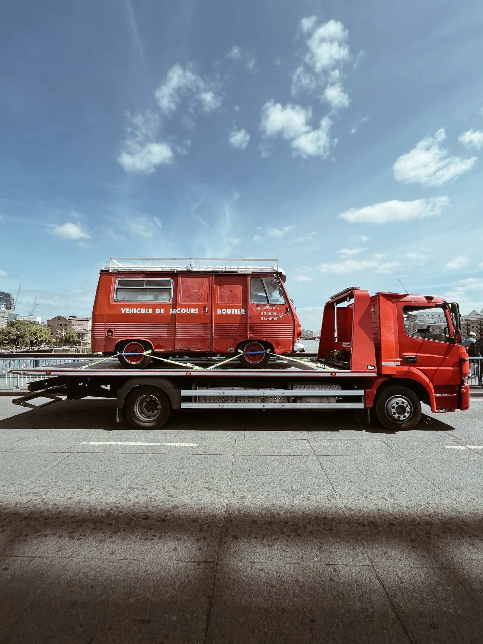 A red flatbed truck carrying a classic van through a city street